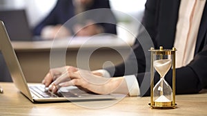 Female working on laptop at office, hourglass measuring time, efficiency at work