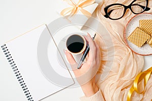 Female workspace with female hand holding coffee cup, notebook, scarf, golden accessories , gift box, waffles, glasses on white