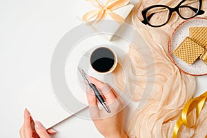 Female workspace with female hands holding card, coffee cup, scarf, golden accessories, ribbon, gift, waffles, glasses on white