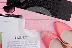 Female workspace with keyboard, sun glasses, shoes, different papers on pink background.