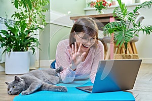 Female on yoga mat waving hand at laptop, in home interior with sleeping cat