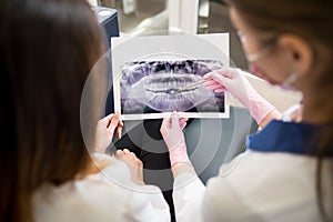 Female young dentist examining x-ray image with female patient in dental clinic and preparing for treatment. Dentistry