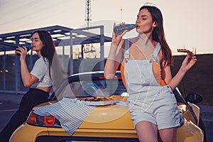 Females drinking carbonated beverage in glass bottles, eating pizza while posing near yellow car on parking lot. Fast