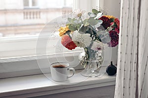 Feminine autumn still life. Cup of coffee, bouquet of colorful dahlia flowers in glass vase on windowsill. Styled moody