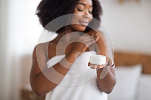 Feminine black woman holding jar of body or face cream, applying skin care product after shower at home