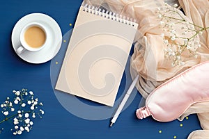 Feminine desk with blank paper notepad, coffee cup, pink sleep eye mask, flowers. Cozy home, happy morning concept. Flat lay, top