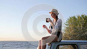 Feminine drinking coffee to old vehicle on coast river, tea ceremony of girl on roof of retro car on nature in sun rays,