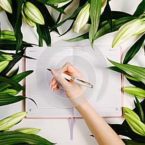Feminine hand writing in notebook surrounded with green fresh flowers on white background. Trendy beautiful workspace