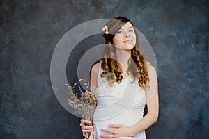 Feminine pregnant young woman in white dress with bouquet of dried flowers touching her belly. Maternity, pregnancy concept.