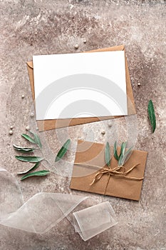 Feminine wedding invitation mockup. Blank card, craft envelope, olive branch and silk ribbon. Beige grunge background. Top view,