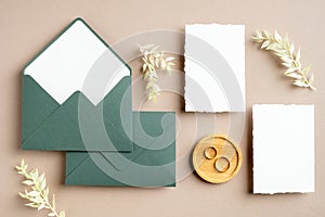 Feminine wedding stationery set. Blank greeting card, green envelopes, dried flowers, golden rings on pastel beige background.