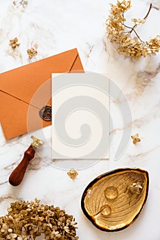 Feminine wedding stationery top view. Blank wedding invitation, bronze envelope, wax stamp, golden rings, dry flowers on marble