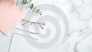 Feminine workspace with glasses, paper notepad, pink notebook, pen, eucalyptus leaf on marble table. Stylish home office desk