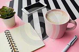 Feminine workspace with smartphone, cup of coffee and blank notepad with copy space on pink background