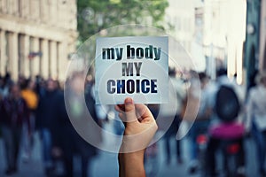 Feminist hands holding a protest banner with the message my body my choice over a crowded street. Human rights concept against