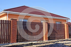 Fence of brown boards and red bricks in front of a private house with a tiled roof