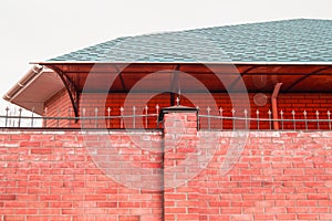 The fence wall is made of red brick, the roof of the house is visible from behind the fence. modern cottage construction