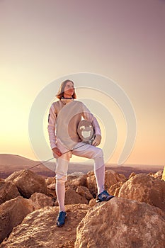 Fencer man standing on top of the rock holding fencing mask and a sword and looking forward seriously