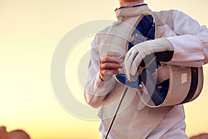 Fencer wearing white fencing costume and holding his fencing mask and a sword on sunny background photo