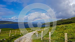 Fences with wooden posts and barbed wire on a dirt trail between the Irish countryside with a rainbow over the Atlantic Ocean