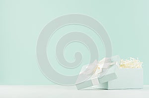 Festive background with opened square gift box with ribbon and knot on soft light green mint menthe wall and white wood table.