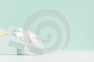 Festive background with opened square gift box with ribbon and knot on soft light green mint menthe wall and white wood table.