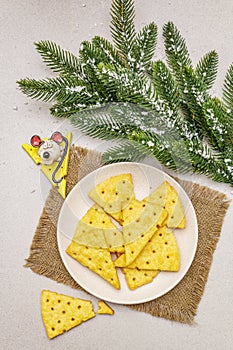 Festive cheese crackers, New Year snack concept. Cookies, mouse figure, fir tree branch, artificial snow, sackcloth napkin. Stone