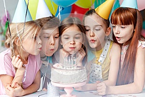 Festive children in party caps, blow candles on delicious cake, make wish, celebrate birthday, have party together, hold
