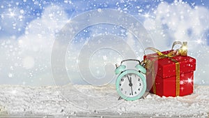 Festive Christmas banner. An alarm clock and a red gift box with a gold ribbon stand on the snow on a blurred blue background with