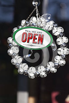 Festive Christmas decorations and open sign with jingle bell wreath hanging in shop store window door. Holiday shopping season.