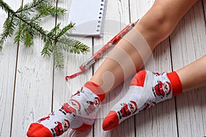 A Christmas photo with feminine feet in bright red socks, a fir twig, a note book and a pencil on a white wooden floor