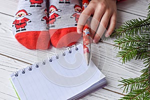 A Christmas photo with feminine feet in bright red socks, a fir twig, a note book and a pencil on a white wooden floor