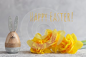 Festive composition with yellow daffodils, Easter bunny, and the inscription Happy Easter against grey wall. Holiday