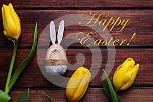Festive composition with yellow tulips, Easter bunny, and the inscription Happy Easter on a rustic wooden background. Holiday