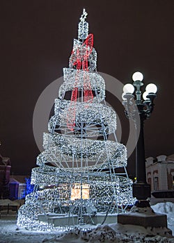 Festive creative Christmas tree made of metal construction with glowing lights of led lamp and city lantern, at night, outdoors