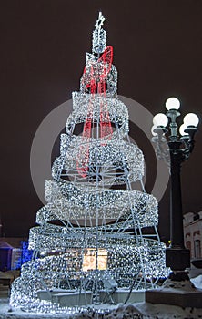 Festive creative Christmas tree made of metal construction with glowing lights of led lamp and city lantern, at night, outdoors
