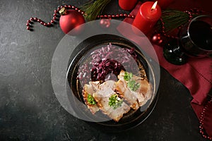Festive dinner, roast pork slices with red cabbage on a dark plate, red candles, wine and Christmas decoration on a dark