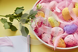 Festive Easter background. Pink bowl with colorful eggs, pink feathers, pink rose and notepad. Happy Easter concept