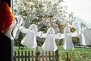 Festive garland with paper ghost shape hanging on the window glass. Halloween holiday home decor concept. Selective