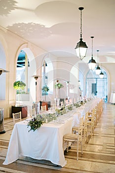 Festive long table with bouquets of flowers in a large hall with glowing chain lamps on the ceiling