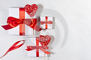 Festive love background - white gift boxes with red silk ribbons, sweet lollipops hearts on white wood board, top view.