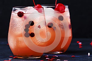 Festive pink cold alcoholic cocktail with red maraschino cherries for Valentines day, two glasses, black background with sweet red