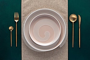 Festive place setting with beige napkin. Empty plates and gold cutlery on dark green background. Top view. Dining table in luxury