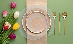 Festive place setting with beige napkin and spring flowers. Empty plates and gold cutlery on green background. Top view. Dining
