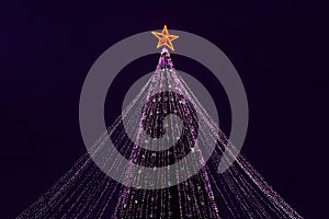 Festive purple xmas lights on big christmas tree with red star in urban central park at holiday evening in winter night