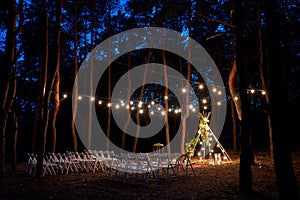 Festive string lights illumination on boho tipi arch decor on outdoor wedding ceremony venue in pine forest at night