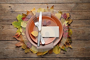 Festive table setting with autumn leaves and blank card on wooden background, flat lay