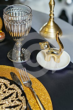 Festive table setting concept. Empty plate, cutlery set, wineglass and golden cat figurine on black table