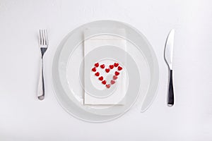 Festive table setting. Empty plate, knife, fork, napkin and red  heart on white table. Top view, flat lay, copy space