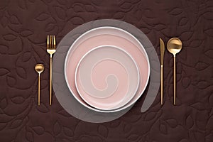 Festive table setting. Golden fork, knife and spoon. Empty ceramic plate with gold cutlery. Dark brown tablecloth. Top view.